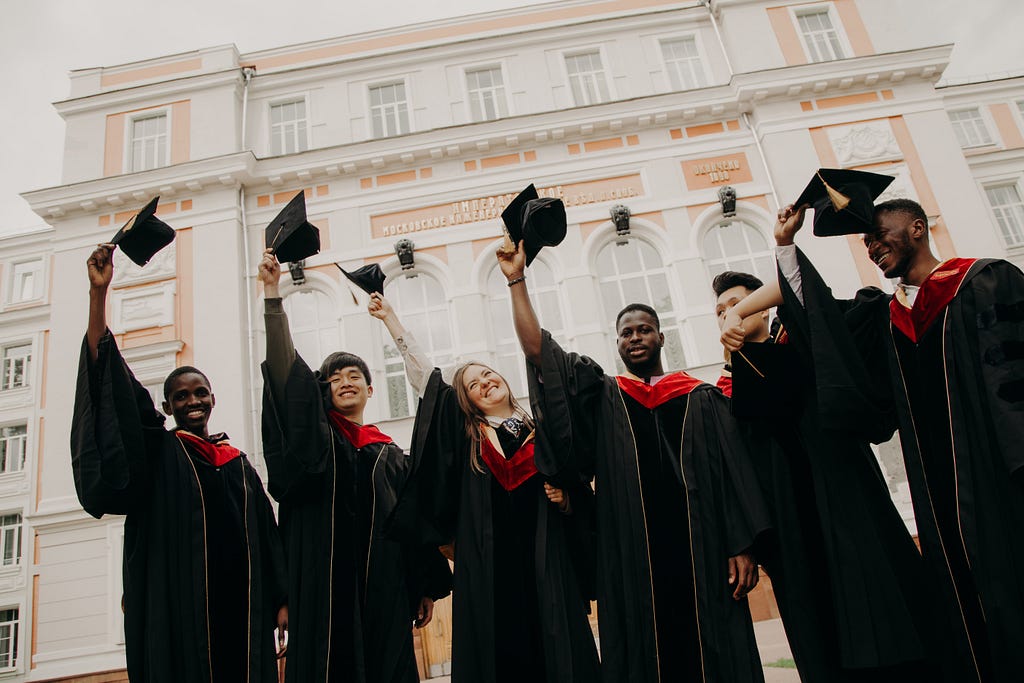 Picture of 6 graduates in tobe, holding their caps in the air.