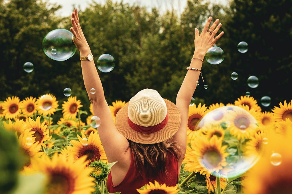 A woman wearing a small sun hat and a red sleeveless shirt throws her hands up in celebration as she frolics in a field of sunflowers that are waist high. Bubbles rain down from the sky.