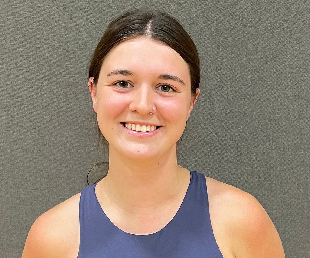 Sarah Gosch stands smilling at the camera in a blue tang top and a gray background behind her.