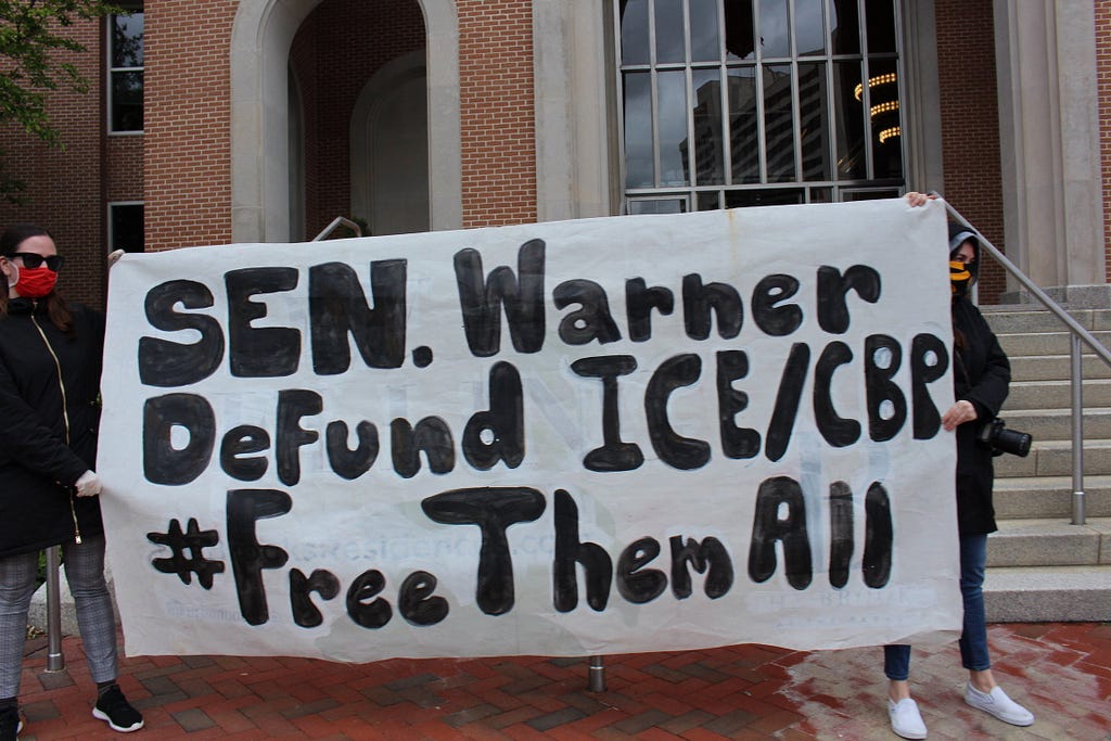 Two people holding a banner that reads “Sen. Warner Defund ICE/CBP #FreeThemAll”.