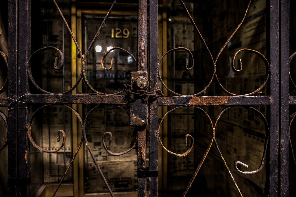Close-up of rusty, wrought-iron gate, locked and held together by a rusty chain. Behind the gate is a double glass door. On the left door, the number 129 sits at top center in gold, outlined in black. The space behind the doors is dark, but two bright lights from the street are reflected in the glass.