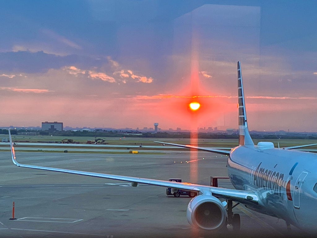 Color photo from inside the Dallas airport looking out onto a perfect pink sunset ‘ball’ and its light rays causing a matching pink ‘cross’ effect appearing in the dusk sky, just about middle and to the left of an American Airlines plane on the tarmac parked at a gate.