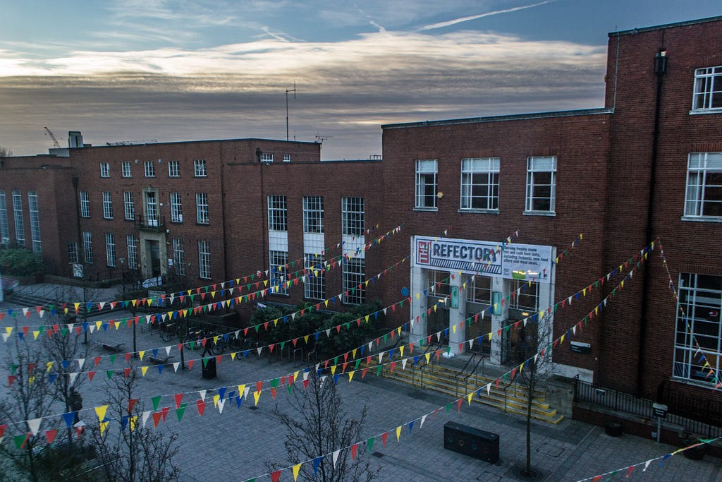 The sun is setting on the Leeds University Union building. Multicolour flags on string hang above the walkway.