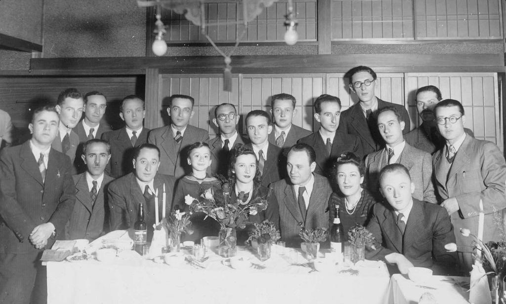 A group of 17 men, two women, and one young boy poses behind a table topped with a white tablecloth and flowers. The adults in the photo are wearing business professional dress.