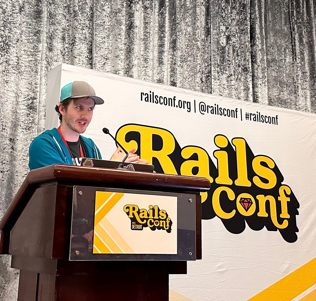 Connor Dudas standing at a lectern, wearing a baseball hat and hoodie. There is a RailsConf banner behind him.