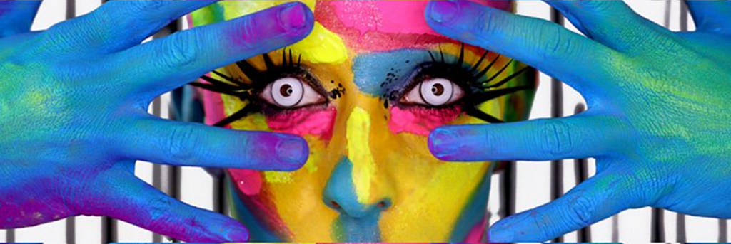 Fotografia de um recorte de rosto de aparência feminina, com as mãos em sua frente e dedos abertos. A figura está pintada com as cores azul, amarelo e rosa.