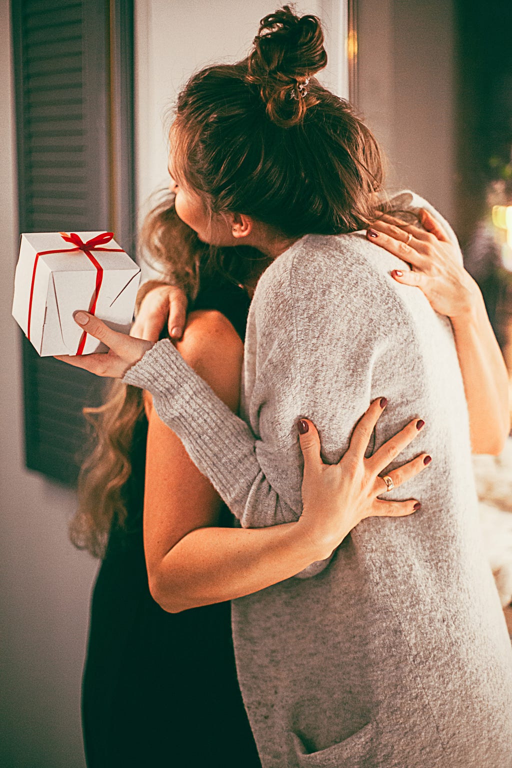 Two women hugging. One woman is holding a small white gift box wrapped in a red ribbon.