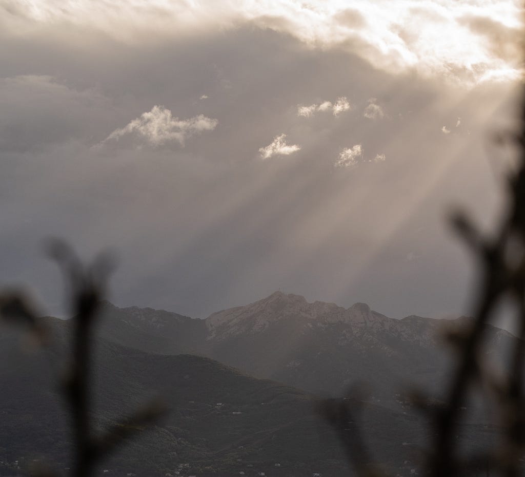 A dark mountain range with light breaking through dark clouds, in the foreground some blurry shapes of branches sticking out from the bottom