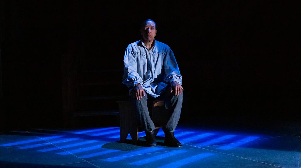 A photograph of a man with brown skin and short dark hair. He wears a light blue shirt with billowing sleeves over gray pants, and sits in a chair alone in a jail cell.