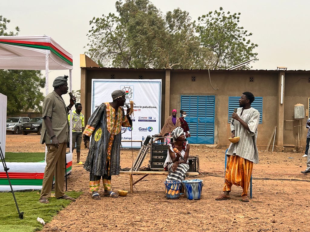 Four actors with microphones stand outside at a launch event for malaria vaccines. The woman is sitting on a bench and has two buckets of water in front of her. The three men are standing and talking to each other.