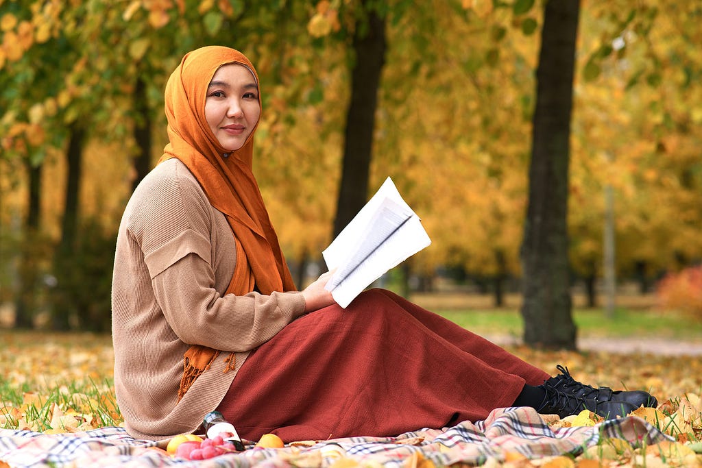 A young female student reader, in hijab scarf, sitting in a golden autumn park and reading the Quran.
