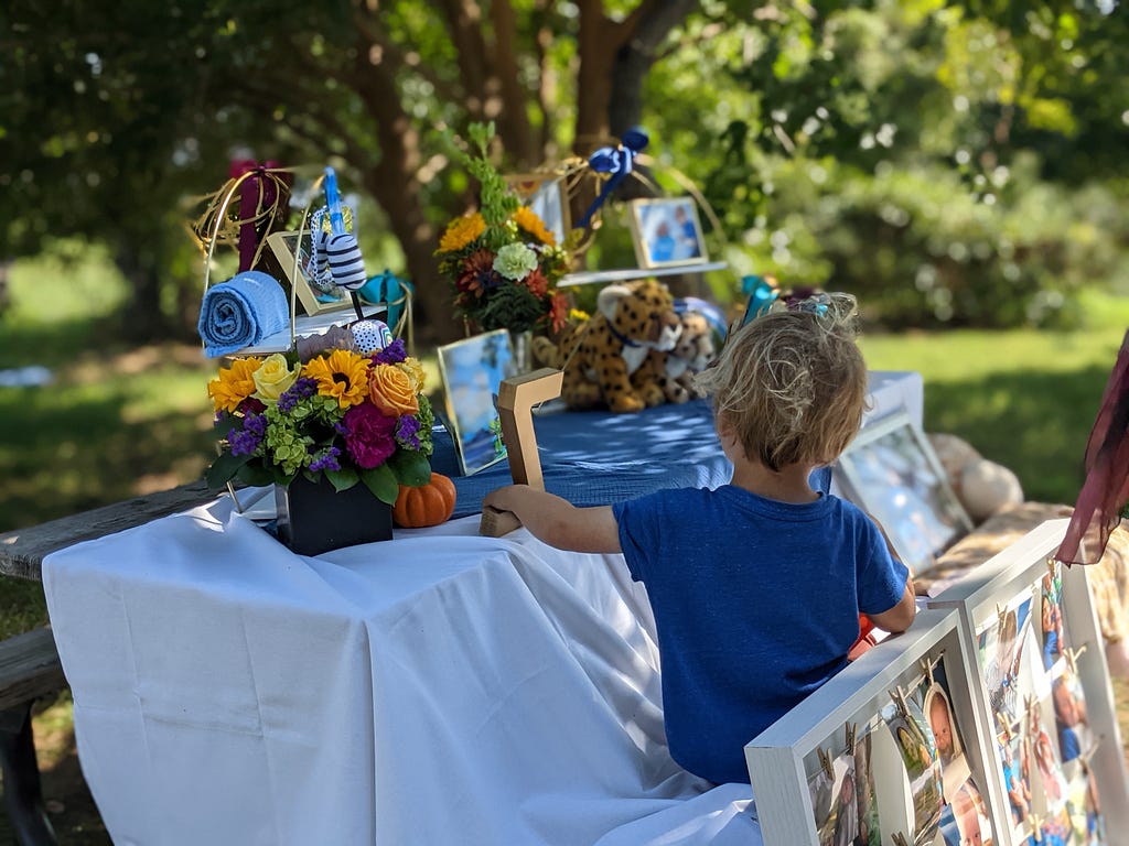 Young child in blue shirt sitting on a table with white tablecloth with photo boards leaning against the table and a display of photos and mementos on the top.