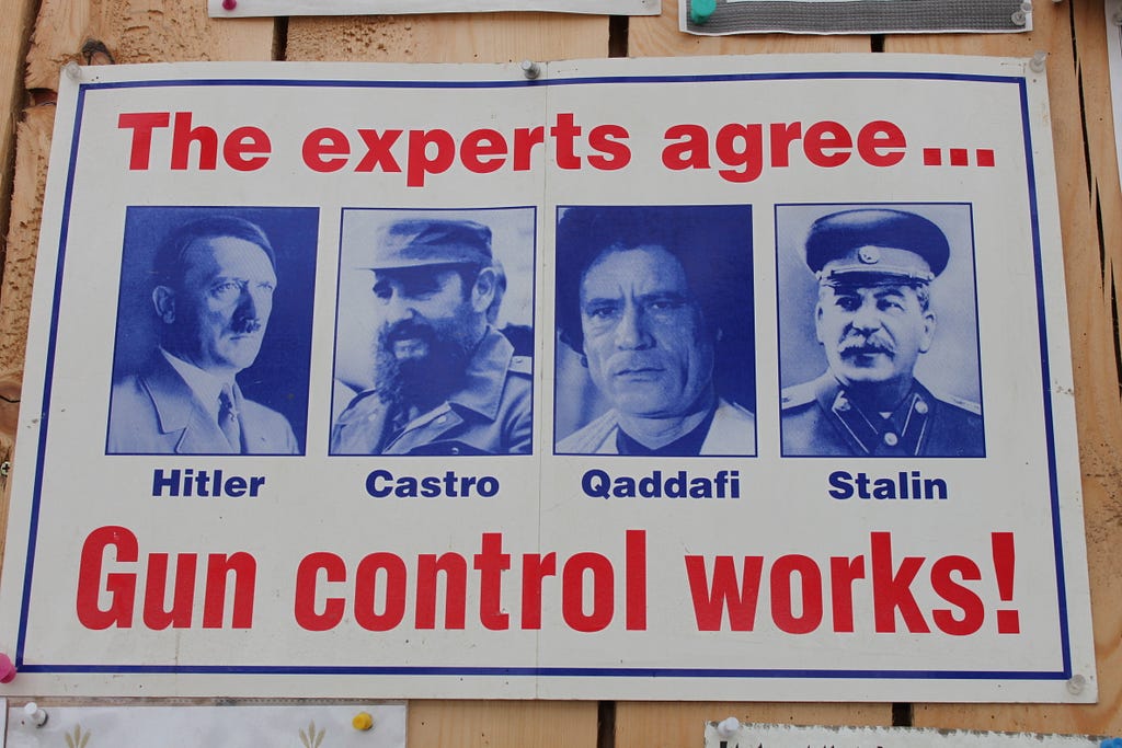 Color photo of a poster pinned to a light wooden wall, of 4 historical dictatatoes in a row in blue headshots left to right: Hitler, Castro, Gaddafi, and Stalin. With bright red text above them stating “The experts agree…” and below them stating in larger red text “Gun control works!”