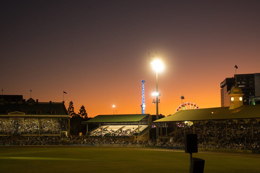 A showground during an orangey sunset with very bright lights illuminating the grass. Multiple stands with shade are in the lower third of the picture.
