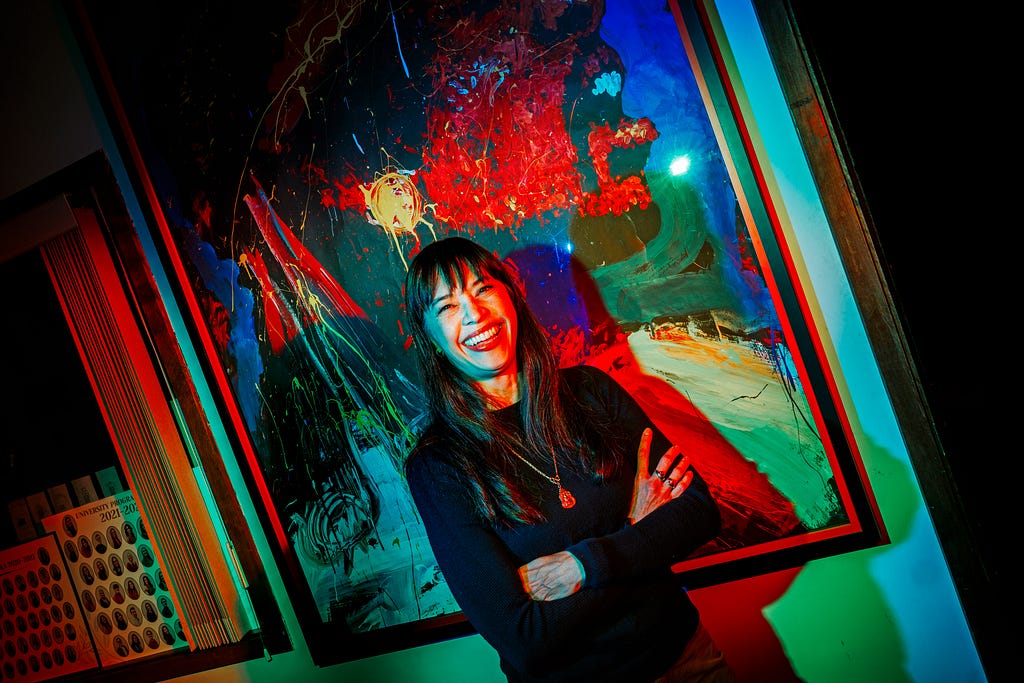 Karen Wills smiles for a photo in front of a painting in the UPC office with blue and red lights shining on her