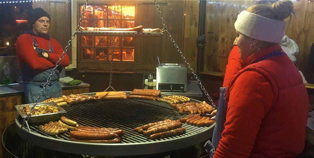 Two people wearing red shirts and overalls watch over a ring of sausages on the grill.