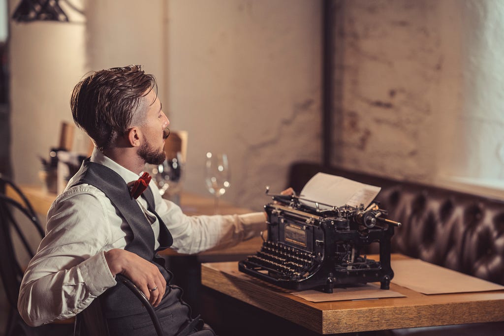 Young, moustachioed man in dress shirt and vest sits at an old-fashioned desk in front of a typewriter.