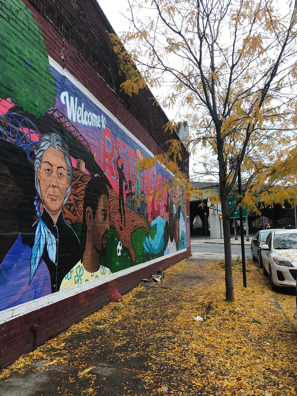 Colorful wall art depicting diverse people lives on a Red Hook building next to black tree with falling yellow leaves