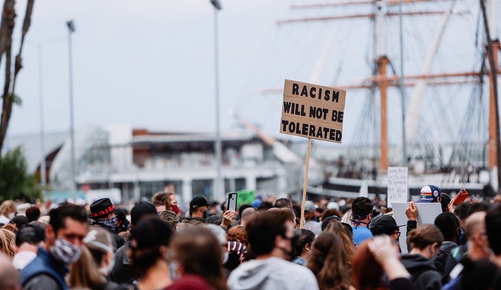 A crowd protesting with a sign held up saying “Racism will not be tolerated”.