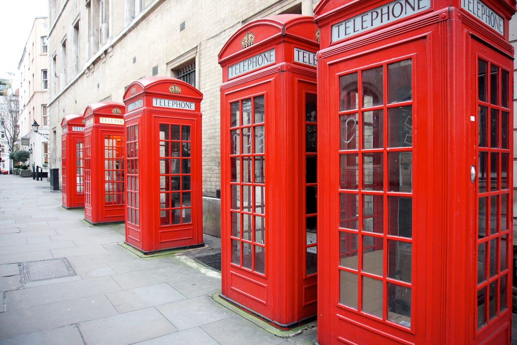 Five, red, British-looking telephone booths on a walkway; adjacent to a wall.