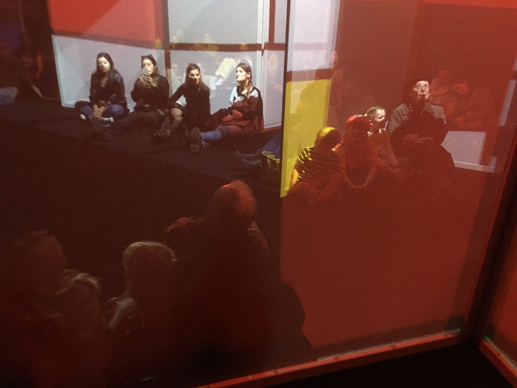 Visitors young and old sit on the floor in a darkened room through which a net screen weaves, on which is projected a motion-graphics interpretation of Piet Mondrian’s “Composition with large red plane, yellow, black, gray and blue”