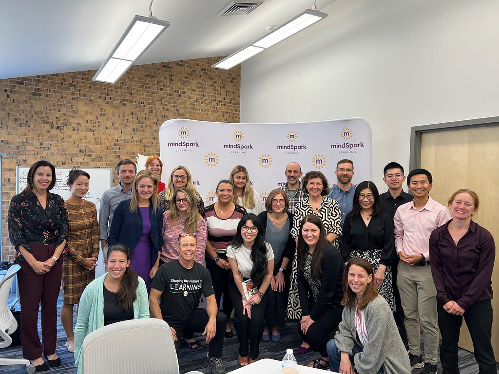 Group of about 20 professionally dressed people inside a brightly lit room smiling for a photo. Behind them is a backdrop that reads “mindspark.”