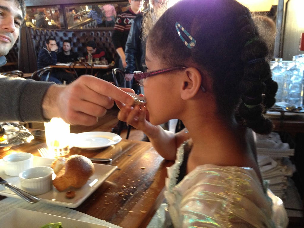 A young girl at a restaurant tries an oyster with help from her dad