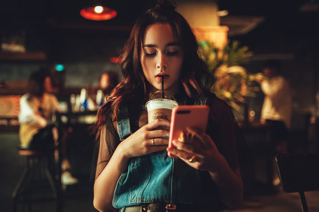 A young woman in a coffee shop looks at her phone while sipping a drink