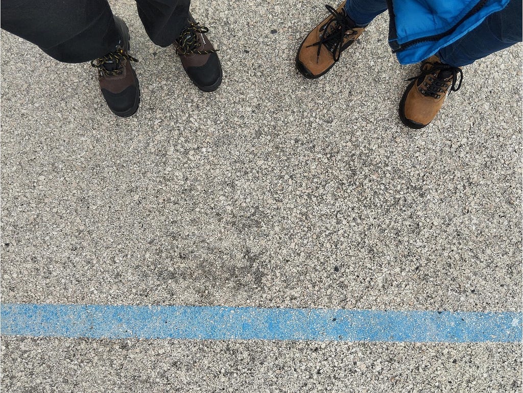 Imagem de chão e pés de duas pessoas com calçado técnico, adequado a ambiente ATEX, junto de uma linha azul demarcada no chão para indicar a zona de passagem autorizada a visitantes.