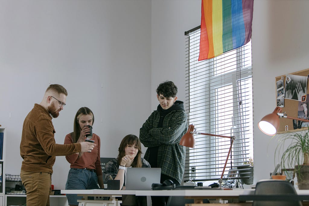 Four people gathered around a laptop in a white office, a window is partly behind a rainbow flag.