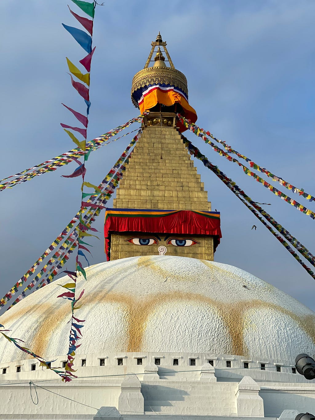 Large Buddhist Stupa (round concrete dome) with the Buddha eyes looking over the top of the dome. The eyes are part of the large gold-colored pyramid rising from the dome and there are many prayer flags strung around the stupa from the top of the pyramid.