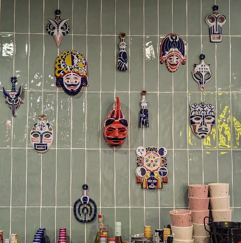 A collection of ceramic masks painted mostly in blues, whites, reds and yellows, hanging above a bar on a tiled grey wall.