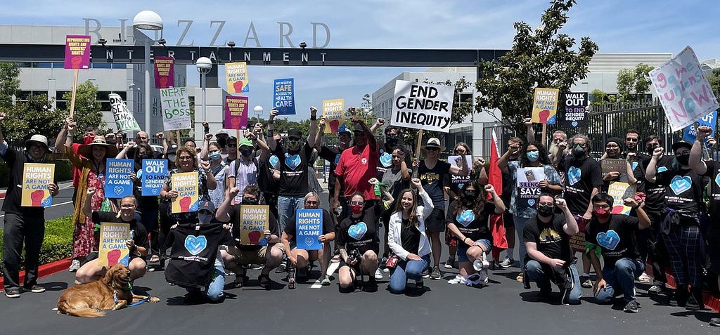 Dozens of Activision Blizzard workers and allies pose in front of the Blizzard Entertainment gates during a walkout. They hold signs that say WALK OUT, END GENDER INEQUITY, HUMAN RIGHTS ARE NOT A GAME, and similar slogans