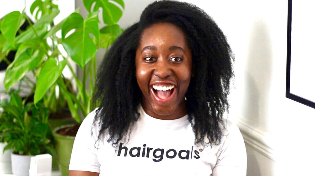 Dr Adaku Jennifer Agwunobi is photographed with a white t-shirt on that reads ‘hairgoals’ and a green potted plant in the background.