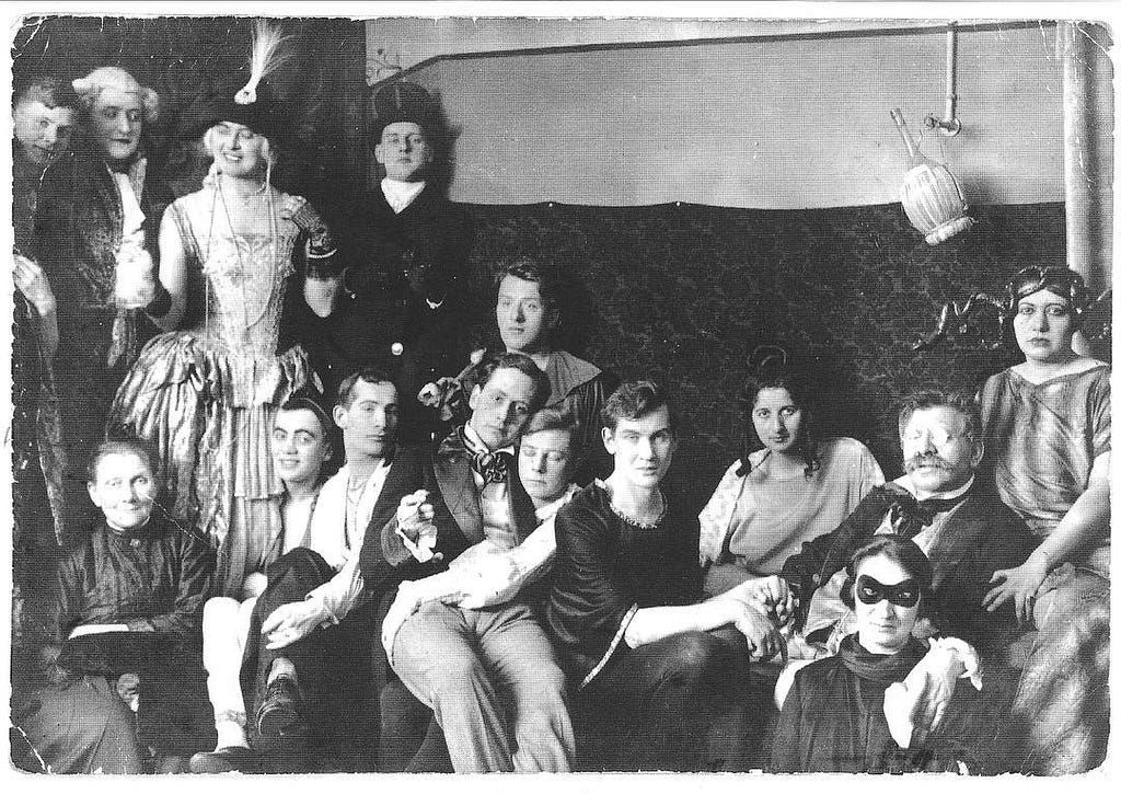 A black and white photo of a group of people in formal wear or costumes. The guests are primarily masculine-presending, but some are in dresses and others are androgynous to the viewer. They mostly look happy, and some are touching each other affectionately.