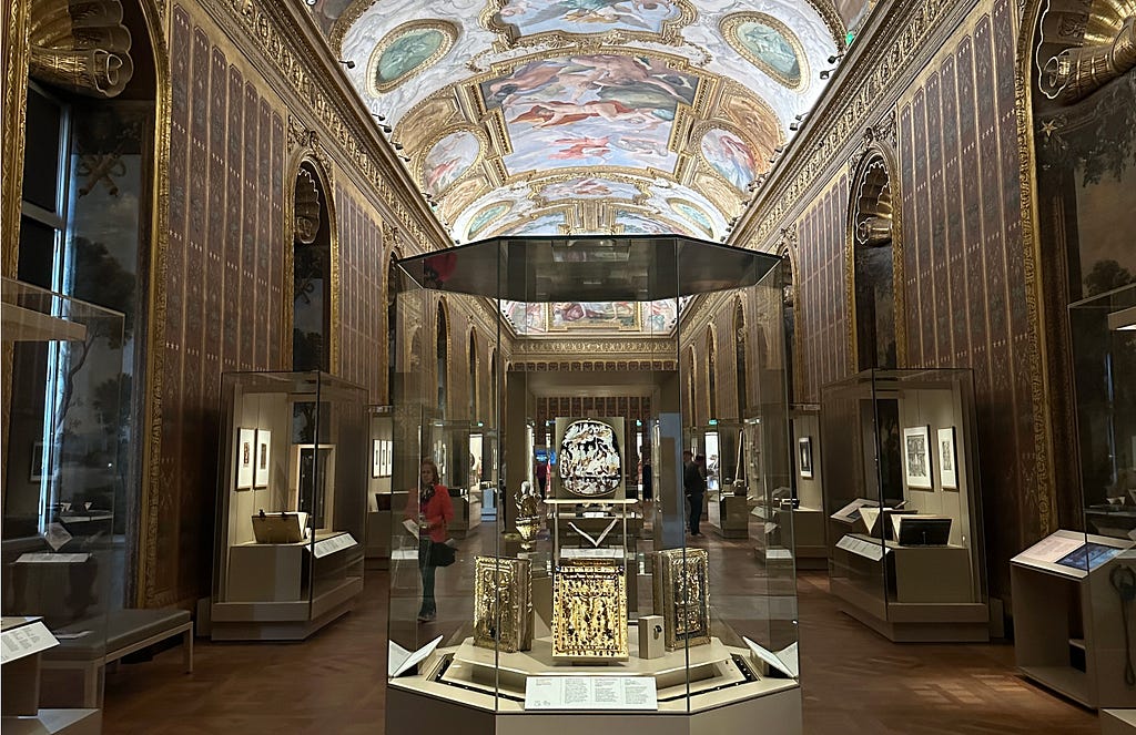 A sumptuous Mazarin gallery (BNF RIchelieu) with a barrel-vaulted ceiling adorned with colorful frescoes, flanked by ornate gilded wall panels and large display cases containing historical objects. The central exhibit features an intricately carved piece of ivory.