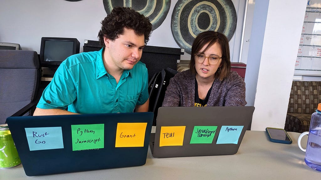 A man and a woman in front of two laptops, both looking at the one to their left, mid-thought as they solve a coding problem.