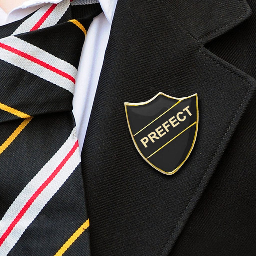 A close-up of a school tie and black school blazer. There is a Prefect badge pinned on the lapel of the blazer.