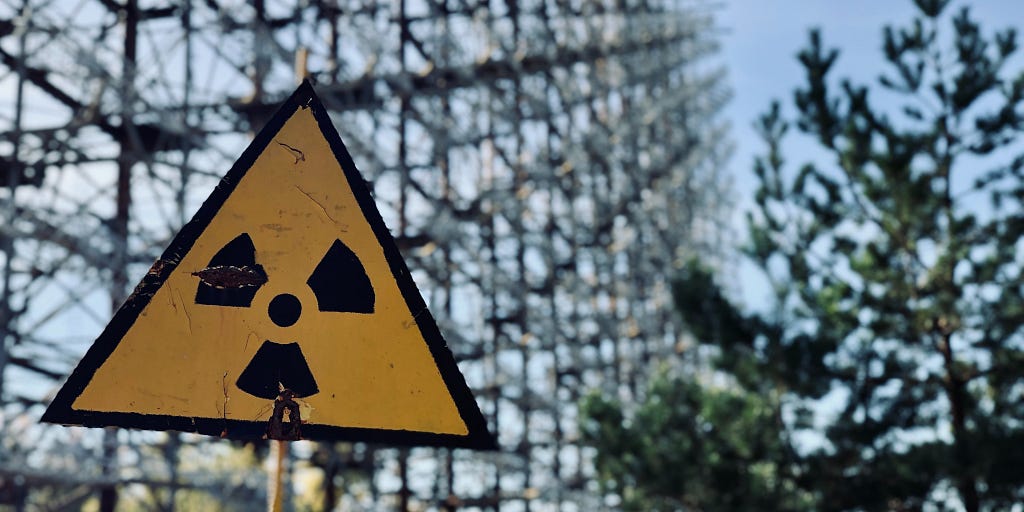 A yellow “radioactive” warning sign in the foreground. Trees and what looks like a fence are out of focus in the background.
