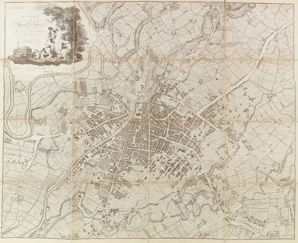 A detailed map of Manchester showing the city surrounded by pastures. A decorative title image adorns the map which includes Manchester personified as a female, a cotton mill, beehive, and the female form of Britannia.