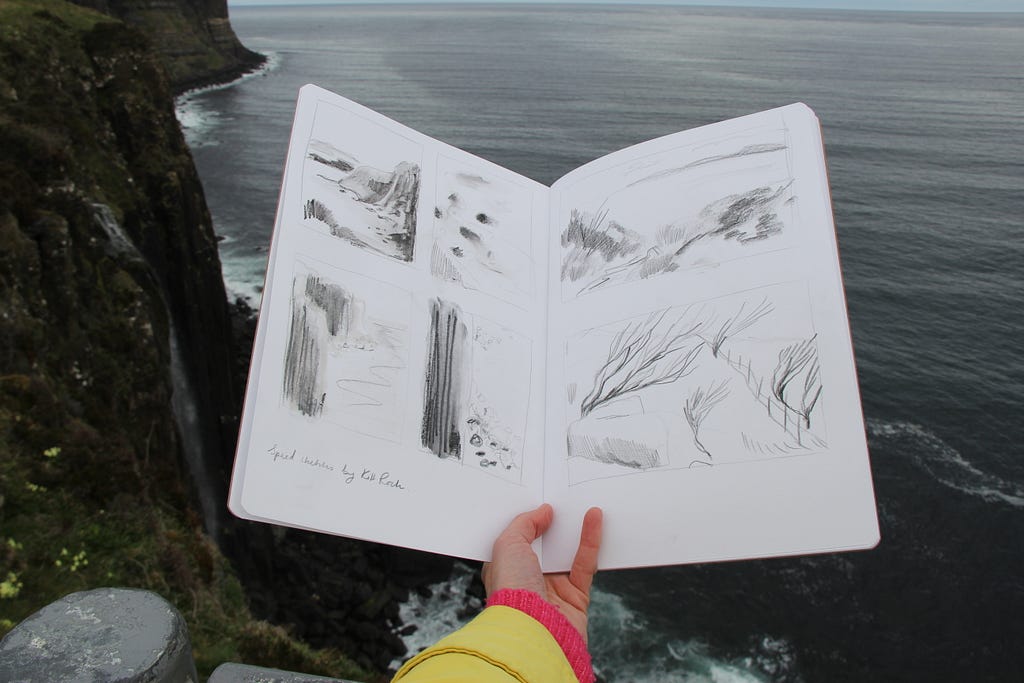 Photo of Helen’s hand holding a work-in-progress sketchbook open in front of an atmospheric coastline