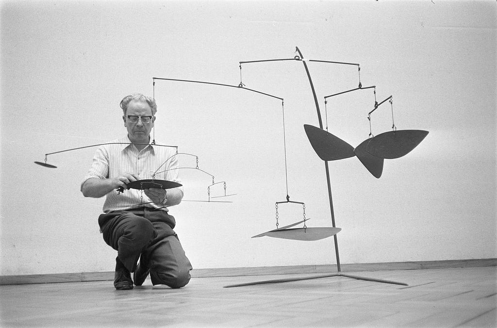 In black and white, against a plain interior background, a middle-aged man in glasses kneels beside a mobile of fan-like metal shapes hanging freely from three short bars attached to a small stand some three or four feet tall.