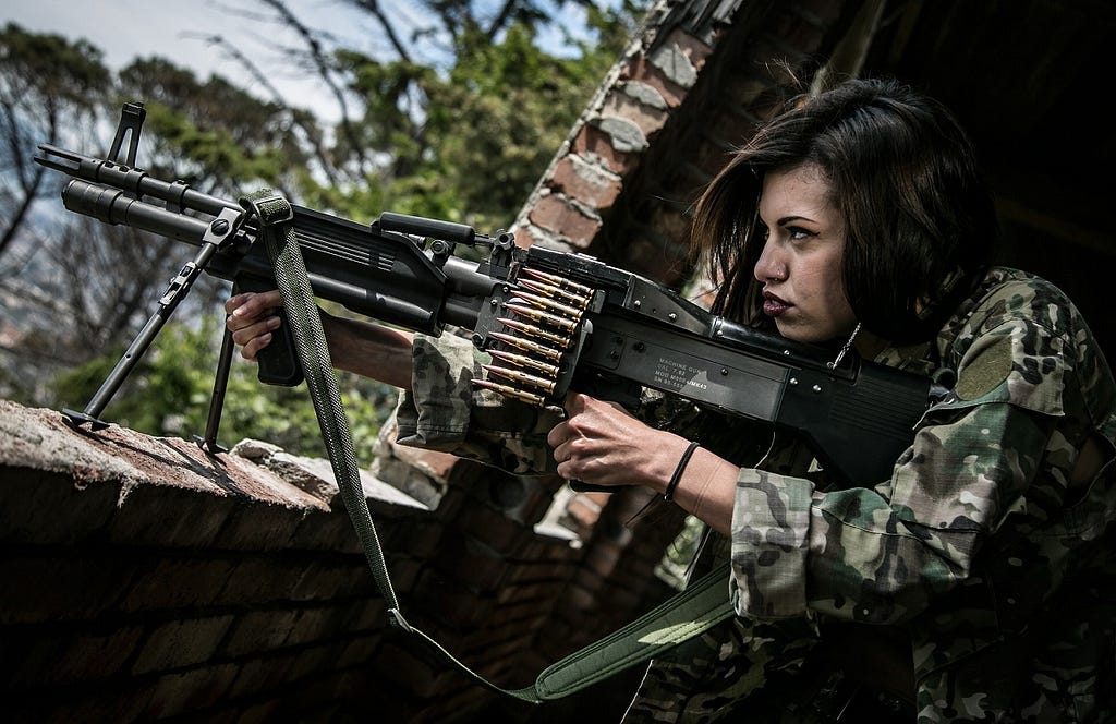 a woman in military fatigues handles a machine gun propped on a brick wall.