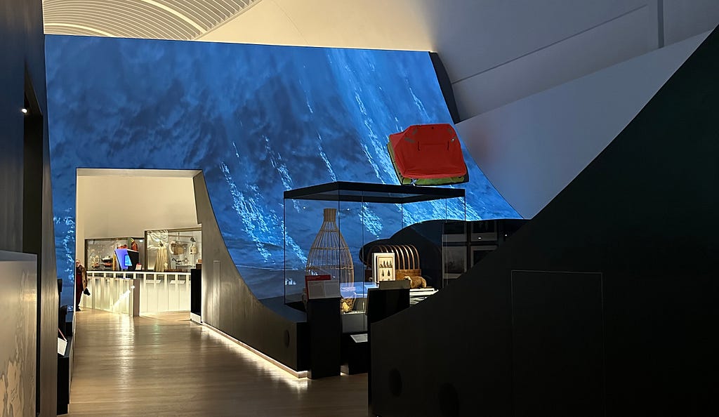 Interior of Musée de la Marine featuring an expansive video mapping of a rolling blue wave as a backdrop. An elevated bright red boat model adds a vivid splash of color to the serene, dark-toned room