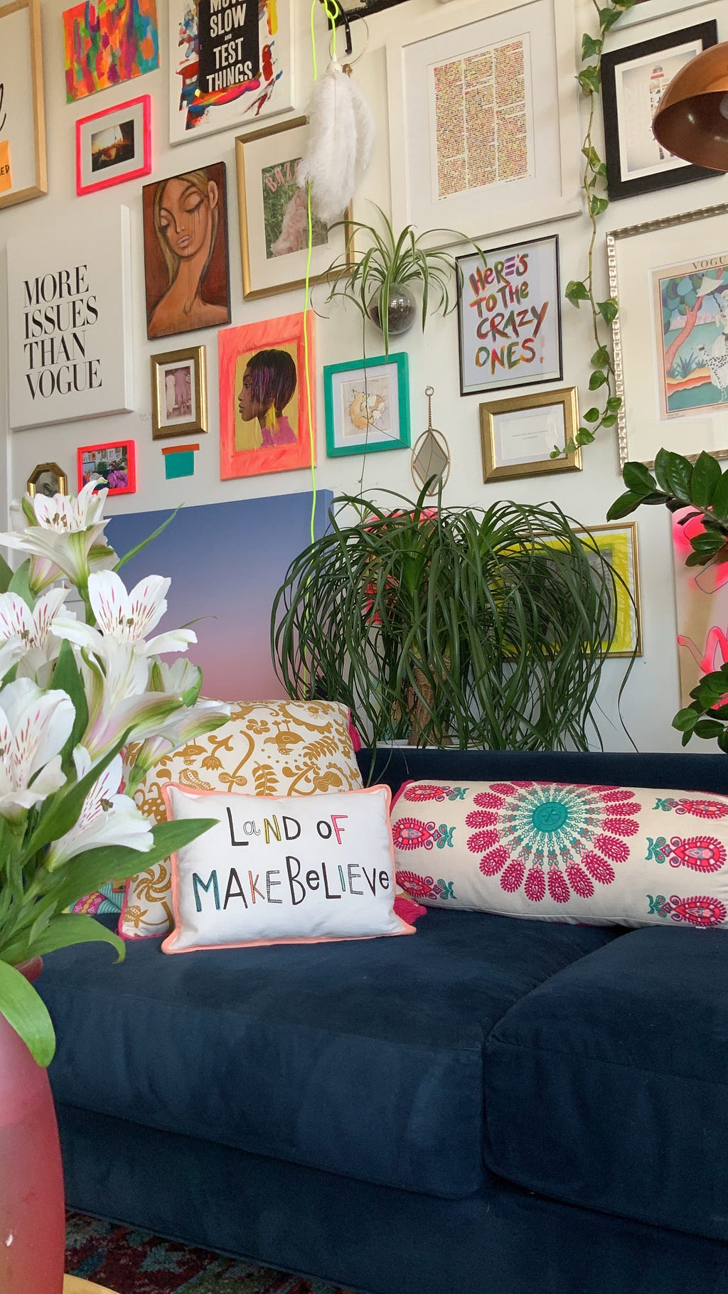 A room features vivid patterns and colors, many framed pieces of art on the wall, and a healthy, robust ponytail palm behind the couch.
