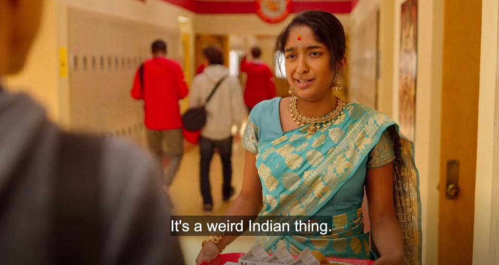 An image of Devi at the school for Ganesh Puja (an Indian celebration of the god Ganesh), where she runs into Paxton (i.e. super hot white-passing guy she’s majorly crushing on). He questions why she is at school on the weekend, to which she answers with the dialogue subtitled “Its a weird Indian thing.”