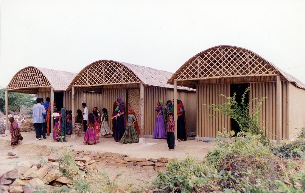 Indian villagers gather in front of three buildings made of paper logs