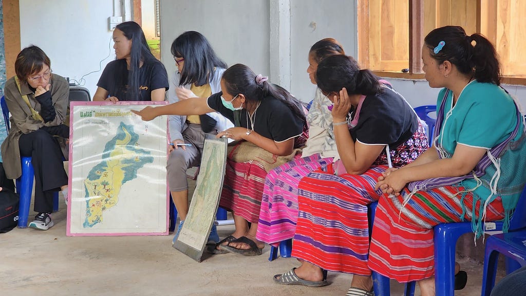 7 women are visible, they are wearing colourful traditional clothing and one of the women is pointing at a map.