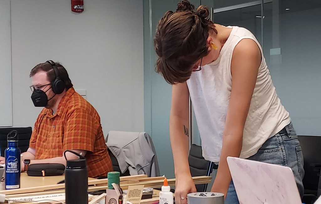 Photo of 2 teachers in an MIT classroom. The teacher on the left is looking at a laptop and wearing headphones. Teacher on the right is working with wood and glue.