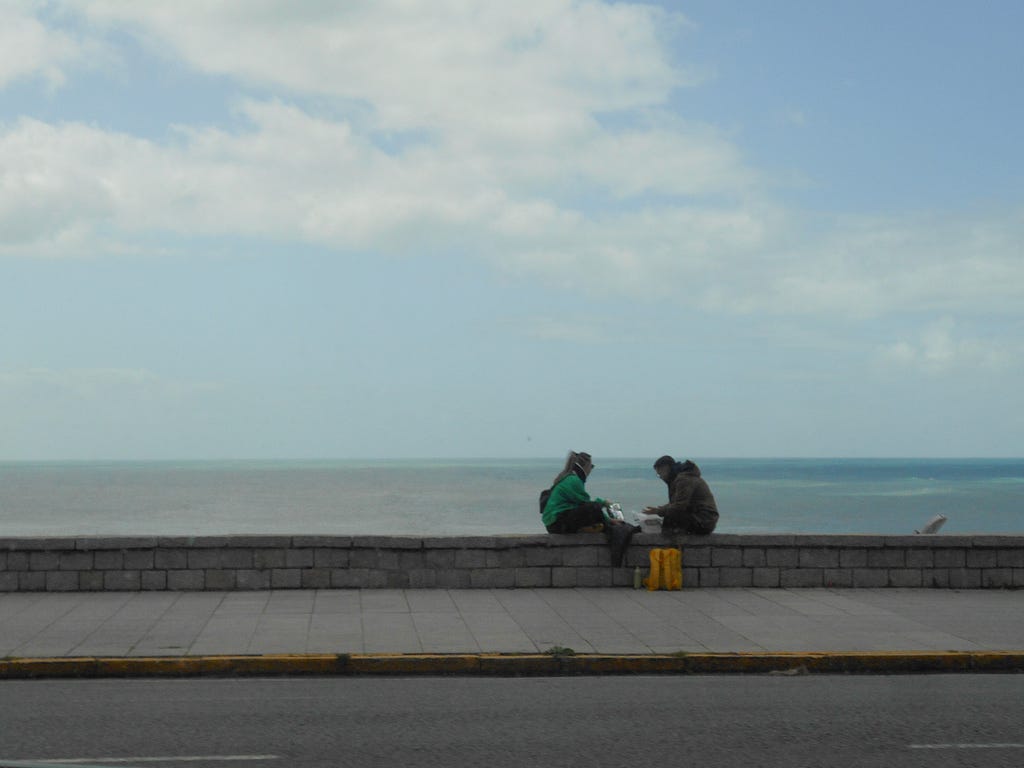 Two people sitting by the sidewalk, facing each other, with the sea behind.